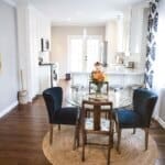 Bright kitchen with white cabinets, a glass table with blue chairs, floral centerpiece, and bird artwork on the wall.