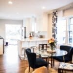 Modern kitchen and dining area with white cabinets, stainless steel appliances, a glass table, blue chairs, and floral curtains.