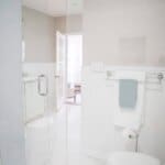 Modern bathroom with glass shower, white tiles, toilet, and towel rack. Open door leads to a bright, adjacent room.